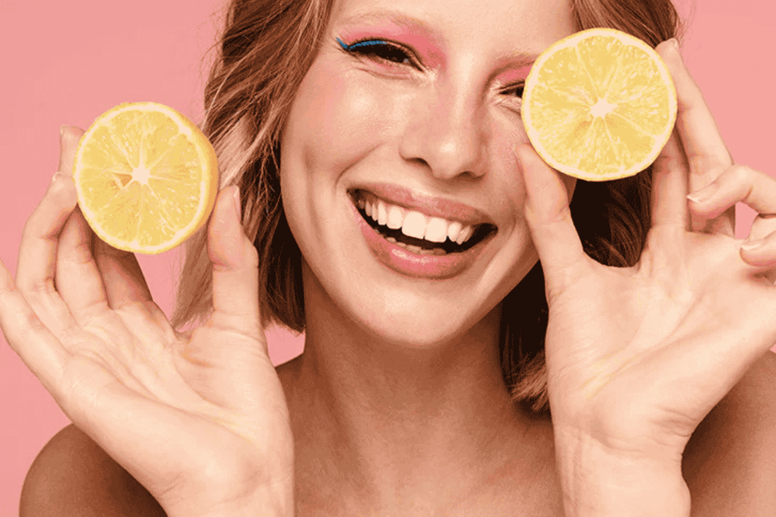Woman holding two orange halves smiling with a pink background.