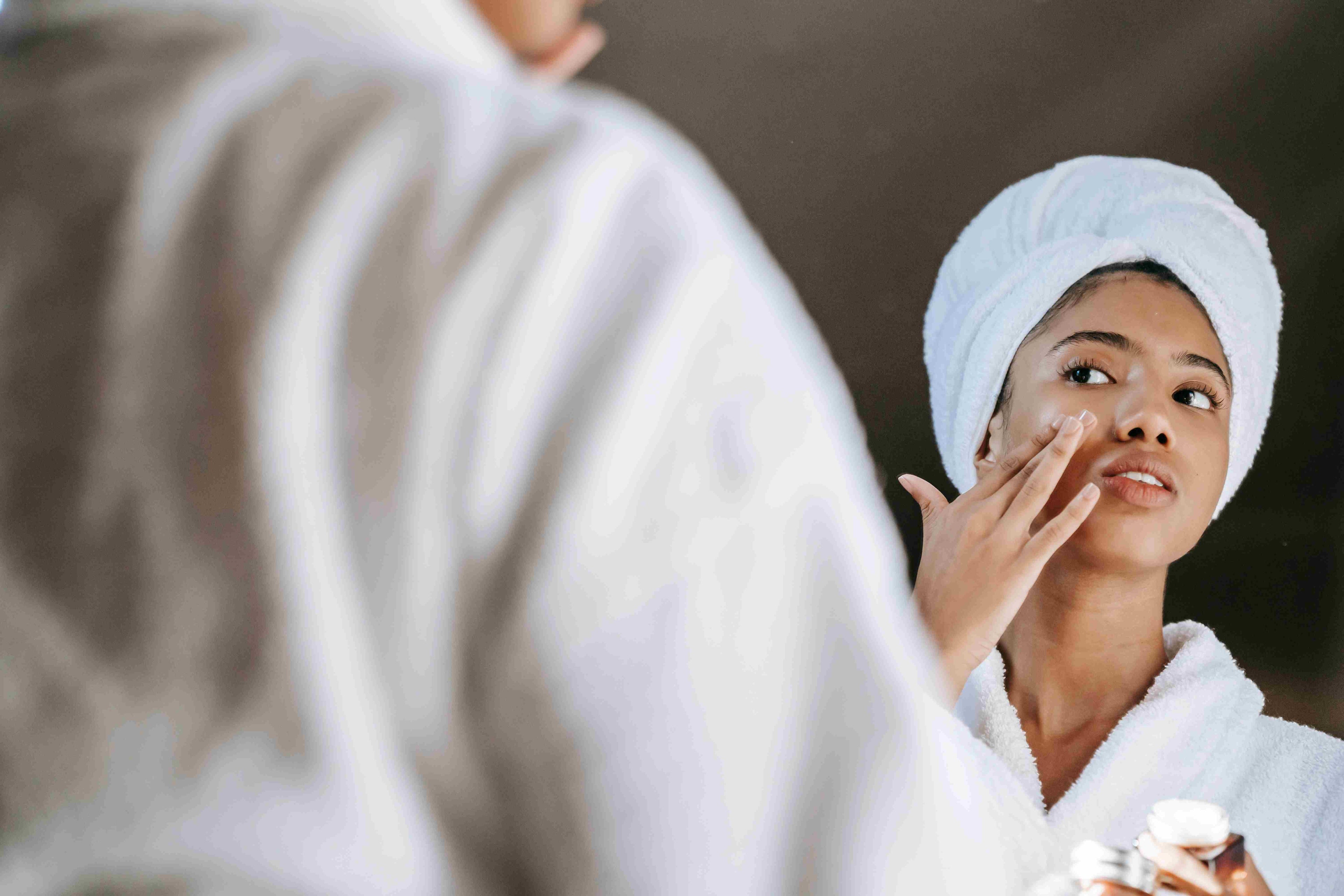 A young woman applies skincare cream while wearing a bathrobe in front of a mirror.  More info  Share
