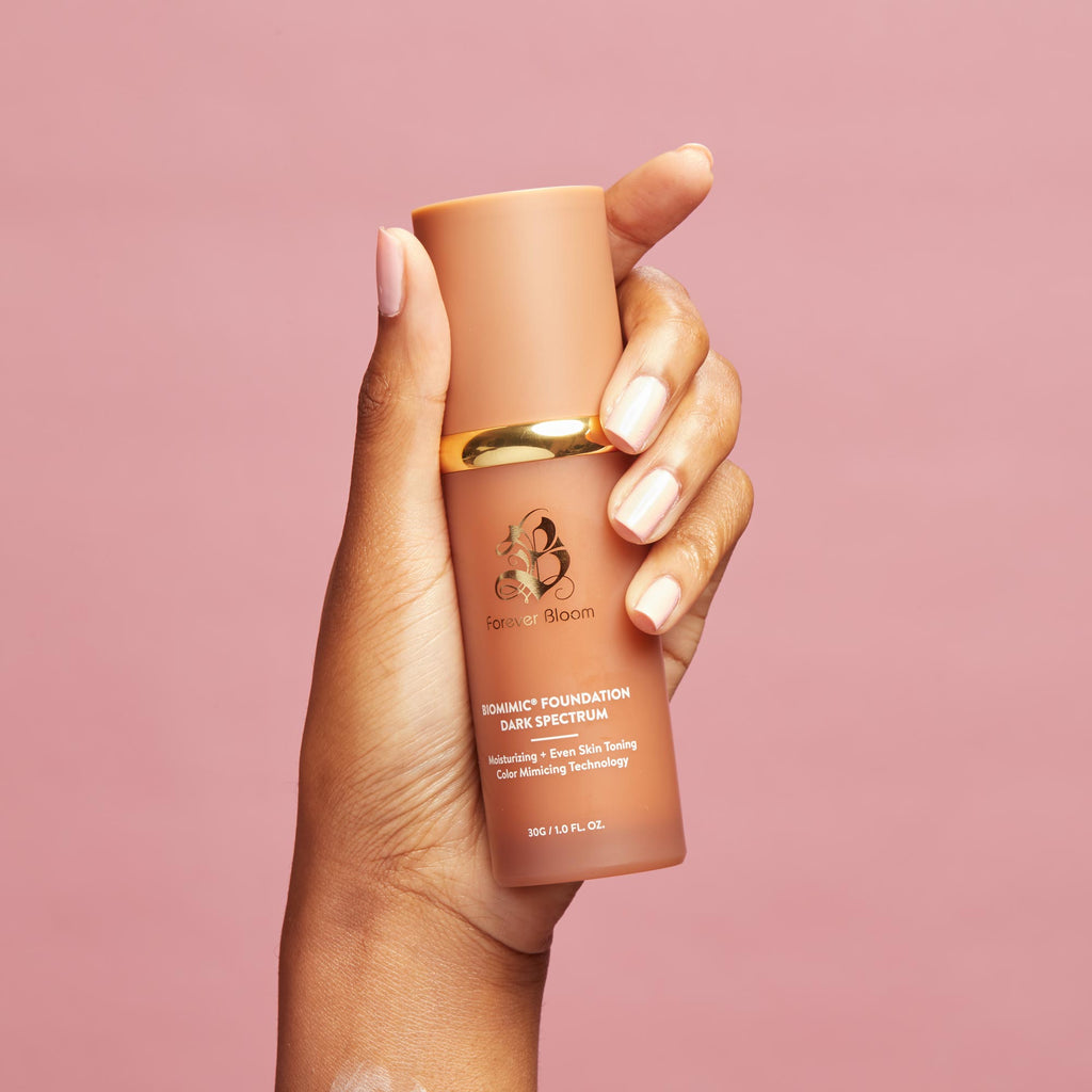 Hand holding a tan-colored biomimic foundation  bottle with a gold cap against a pink background