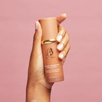 Hand holding a tan-colored biomimic foundation  bottle with a gold cap against a pink background